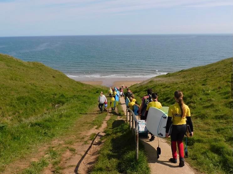 group of people with surfboards walking down towards cayton bay